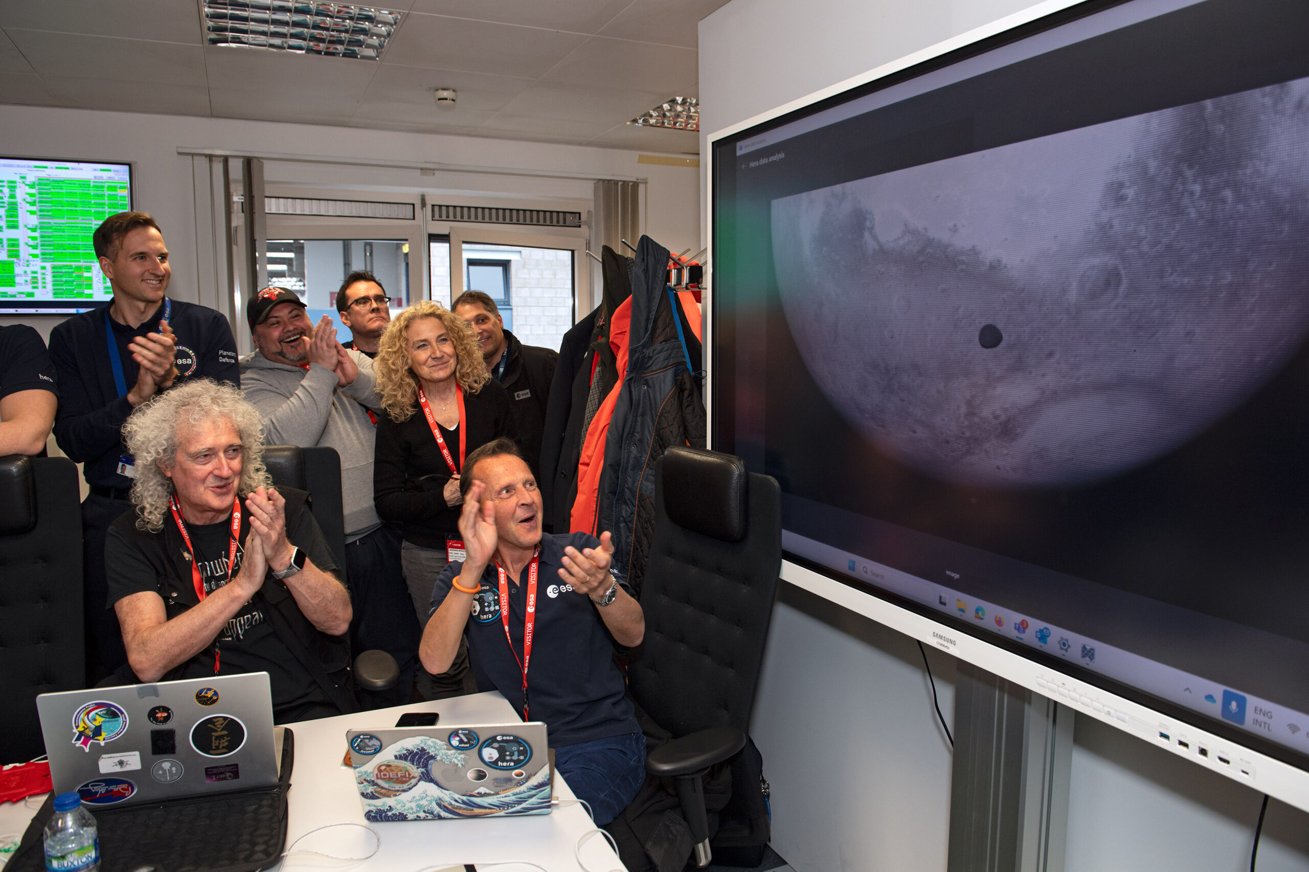 Un grupo de científicos e ingenieros de la ESA celebra frente a una pantalla grande que muestra una imagen en blanco y negro de Marte con su luna Deimos cruzando su superficie